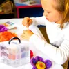 A young girl with Down syndrome is engaged in a craft activity, likely felting, using colorful materials and tools laid out on a table. Produktbild-Vorschau