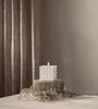 A white bubble candle with a lit LED wick sits on a stone base, surrounded by dried plants and eucalyptus, with a textured curtain in the background. Podgląd zdjęcia produktu