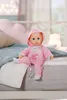 A baby doll dressed in pink pajamas sits on a bed with pillows and a blanket. Náhled obrázku produktu
