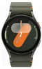 A close-up, front-facing view of a green Samsung Galaxy smartwatch with a dark gray bezel and strap. The watch face displays a colorful, abstract design with orange and gray shapes, and white hands indicating the time. Small blue and orange accents are vis Produktbild-Vorschau