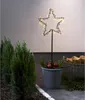A star-shaped LED garden stake with warm white lights is placed in a pot with greenery and red apples, next to a basket of firewood and small evergreen trees. Podgląd zdjęcia produktu