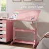 A pink adjustable children's desk with drawings and art supplies on it, next to a pink drawer unit. The scene is set in a child's room. Náhled obrázku produktu