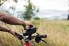 A person's hands are shown adjusting a black wedge case mounted on the handlebars of a red bicycle. The background is a blurred natural landscape with tall dry grass and distant hills under an overcast sky. Anteprima dell’immagine prodotto