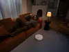 A man is relaxing on an orange couch while a robot vacuum cleaner operates on the floor. Produktbild-Vorschau