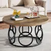 A round coffee table with a wood-grain top and a black metal base with circular accents, placed in a living room setting with a sofa in the background. Podgląd zdjęcia produktu