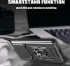A close-up of a black and silver phone case with a ring holder, placed on a textured surface, with a blurred laptop and headphones in the background. Produktbild-Vorschau