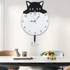 A cat-shaped wall clock with a pendulum shaped like a fish skeleton hangs on a white wall above a dining table. Produktbild-Vorschau