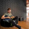 A man is playing a guitar while sitting on a bean bag chair in a room with a brick wall. Náhľad obrázku produktu