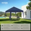 A navy blue pop-up gazebo is set up on a green lawn in a backyard setting, with two chairs and a small table placed underneath it. A swimming pool and lounge chairs are visible in the background, along with a modern white house. Náhled obrázku produktu