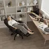 A woman is relaxing in a black ergonomic office chair in front of a bookshelf and a desk with a computer. Anteprima dell’immagine prodotto