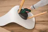 A person is using a cordless screwdriver to attach a wooden leg to the underside of a white chair. Produktbild-Vorschau