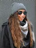 A woman wearing a beanie, sunglasses, and scarf stands in front of a textured metal wall. Produktbild-Vorschau
