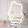 A vanity mirror with LED lighting sits on a marble-like surface next to a pink makeup case and some makeup items. The mirror reflects a bedroom scene with a bed and artwork on the wall. Produktbild-Vorschau