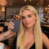 A woman is holding up a phone case in a cafe setting. Produktbild-Vorschau