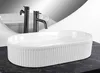 A white oval countertop sink with a fluted exterior sits on a dark countertop, with two dark faucets behind it and a curved mirror reflecting the scene. Anteprima dell’immagine prodotto