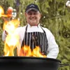 A man in a chef's hat and apron is grilling food over a large flame in a garden setting. Produktbild-Vorschau