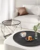 A coffee cup, cookies, and a plant are arranged on a dark round tray on a light-colored ottoman. A black metal coffee table with a glass top is visible in the background. Náhled obrázku produktu