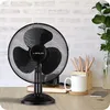A black desk fan is placed on a wooden desk in front of a brick wall. Beside the fan, there is a white mug, a stack of books, and a vase with greenery. Anteprima dell’immagine prodotto
