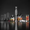 A collection of kitchen appliances including an immersion blender, whisk attachments, and food processor bowls filled with fruit, arranged on a reflective black surface against a dark gray background. Produktbild-Vorschau