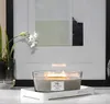 A lit WoodWick candle sits on a white marble tray in front of a framed abstract painting and a plant with white flowers. Podgląd zdjęcia produktu