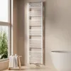 A white towel radiator is mounted on a light beige wall next to a window with greenery visible outside. A small wooden stool with a folded towel and two cups sits in front of the window. Podgląd zdjęcia produktu