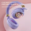 A pair of purple headphones with gold accents, featuring a cartoon cat design on one of the earcups, set against a pink and white background. Voir les images du produit