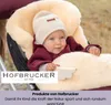 A baby is nestled in a fluffy, beige footmuff inside a stroller, wearing a burgundy hooded cape and a cream-colored beanie. Produktbild-Vorschau