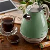 A vintage-style green electric kettle is shown on a wooden table, next to a cup of coffee, a small bowl of coffee grounds, and some nuts. There are also some packaged snacks in the foreground and a tablet and plant in the background. Produktbild-Vorschau