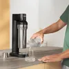 A person is pouring water from a bottle into a glass next to a soda maker. Produktbild-Vorschau