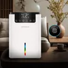 A white dehumidifier is placed on a wooden table in a living room setting, next to a vase with flowers and a cup of coffee. Produktbild-Vorschau