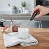 A hand holds a digital thermometer over a glass of milk next to a jar of cocoa powder. Anteprima dell’immagine prodotto