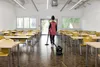 A person is vacuuming a classroom with rows of desks and chairs. Voir les images du produit