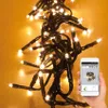 A hand holding a smartphone displaying an app interface, with a string of warm white Christmas lights in the background. Produktbild-Vorschau