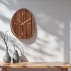A wooden wall clock with gold hands is mounted on a white wall, casting shadows. Two grey vases and a branch are on a wooden table below. Voir les images du produit