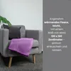 A purple fleece blanket is draped over a gray armchair, next to a dark gray pillow and a green plant leaf. Text describing the blanket's features is overlaid on a gray background on the right side of the image. Produktbild-Vorschau