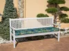 A white garden bench with a patterned cushion sits on a brick patio, next to a tall evergreen tree and a decorative tree. Náhled obrázku produktu