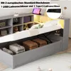 A grey hydraulic daybed with storage, showing the bed lifted to reveal storage compartments filled with folded blankets and pillows. Shelving units are visible above the bed, with decorative items and a tablet charging. Anteprima dell’immagine prodotto