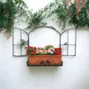 A decorative metal window frame with a flower box filled with red and pink artificial flowers, mounted on a textured white wall with greenery hanging above. Produktbild-Vorschau