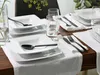 A table setting with white square plates, black cutlery, and glasses of water. Náhľad obrázku produktu