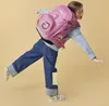 A young girl wearing a pink backpack and jeans is balancing on one leg against a light yellow background. Náhled obrázku produktu