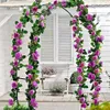A metal pergola arch covered in pink roses and green leaves, set against a white building with shutters. Náhľad obrázku produktu