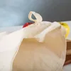 A close-up view of the top opening of a beige fabric bag, with a red apple and a yellow apple blurred in the background. Produktbild-Vorschau