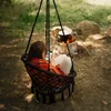 A person sits in a hanging chair, looking at their phone, with a campfire and pot in the background. Voir les images du produit
