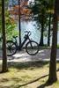 An e-bike parked on a paved path next to a body of water, with trees in the foreground and background. Produktbild-Vorschau