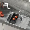 A stainless steel kitchen sink with a black colander and tomatoes inside, next to whole and sliced tomatoes on a countertop. Produktbild-Vorschau
