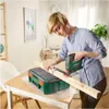 A woman is using a jigsaw to cut a wooden plank that is clamped to a green toolbox which is set up as a workbench. Anteprima dell’immagine prodotto
