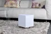 A white cube-shaped humidifier sits on a fluffy rug in front of a white couch with patterned pillows. Produktbild-Vorschau