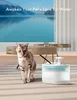 A cat is sitting on a wooden floor next to a pet water fountain, with a large window showing a scenic view in the background. Produktbild-Vorschau