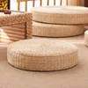 A round, woven straw floor cushion is shown in the foreground, with two more stacked cushions in the background. A woven storage box is visible on the left. Produktbild-Vorschau