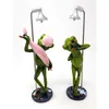Two frog figurines are depicted in a shower setting, each with a shower head above them. One frog is holding a pink towel and wearing a pink shower cap, while the other frog is reaching up towards the shower head. Produktbild-Vorschau